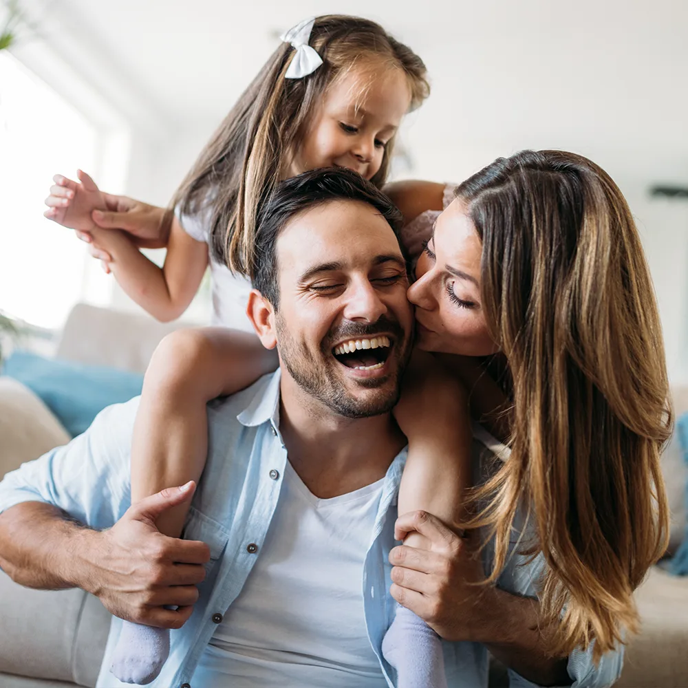 A child on a dad's shoulders while a mother kisses his cheek